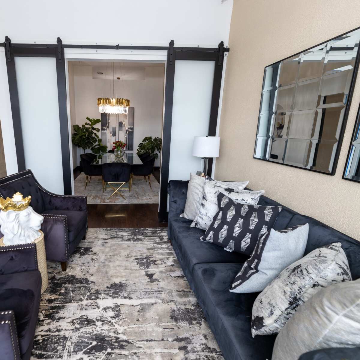 The scene features a stylish living area with two armchairs and a couch, adorned with decorative pillows, leading to a room with a chandelier and plants behind glass sliding doors.