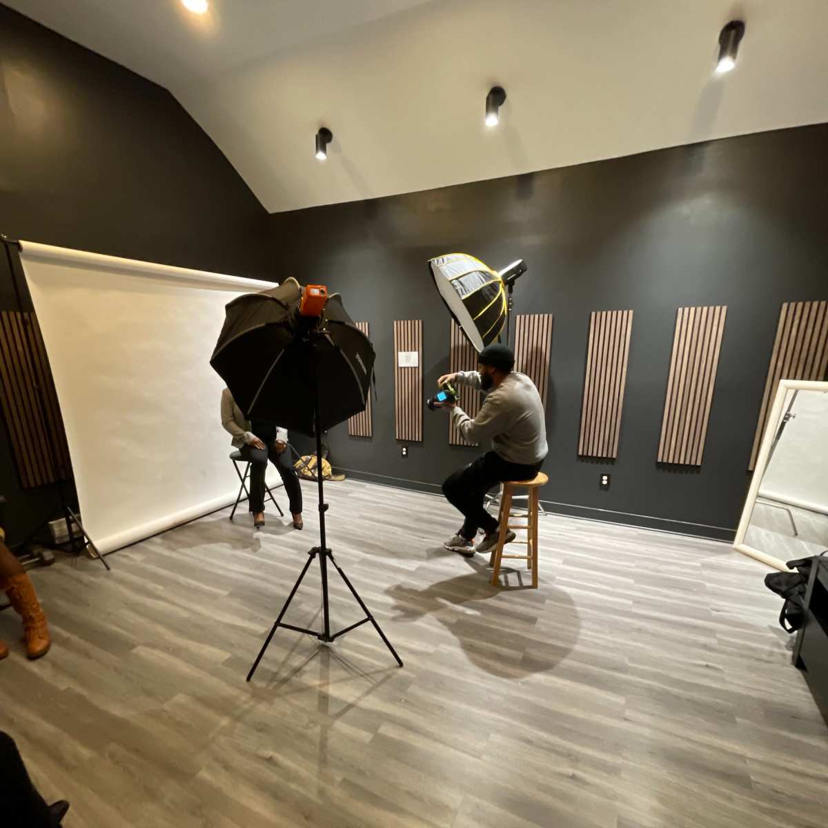 The scene shows a photography studio with a backdrop setup, lighting equipment, and two individuals seated for a photoshoot.