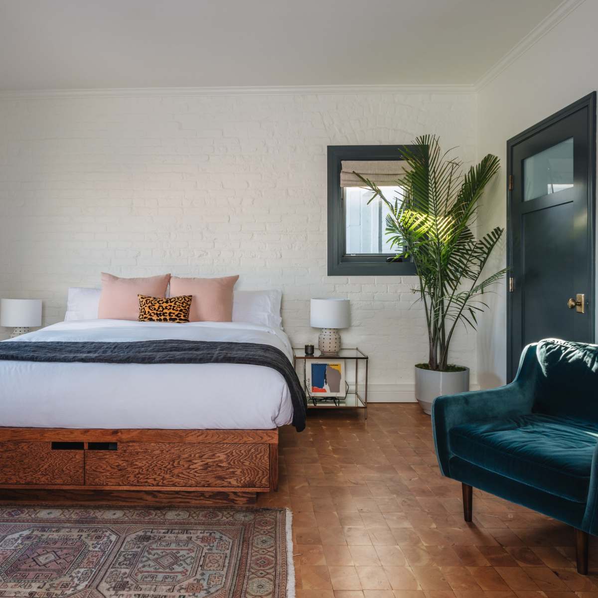 A clean bedroom features a large bed with white bedding, a potted plant in the corner, and a blue armchair beside a wooden nightstand.