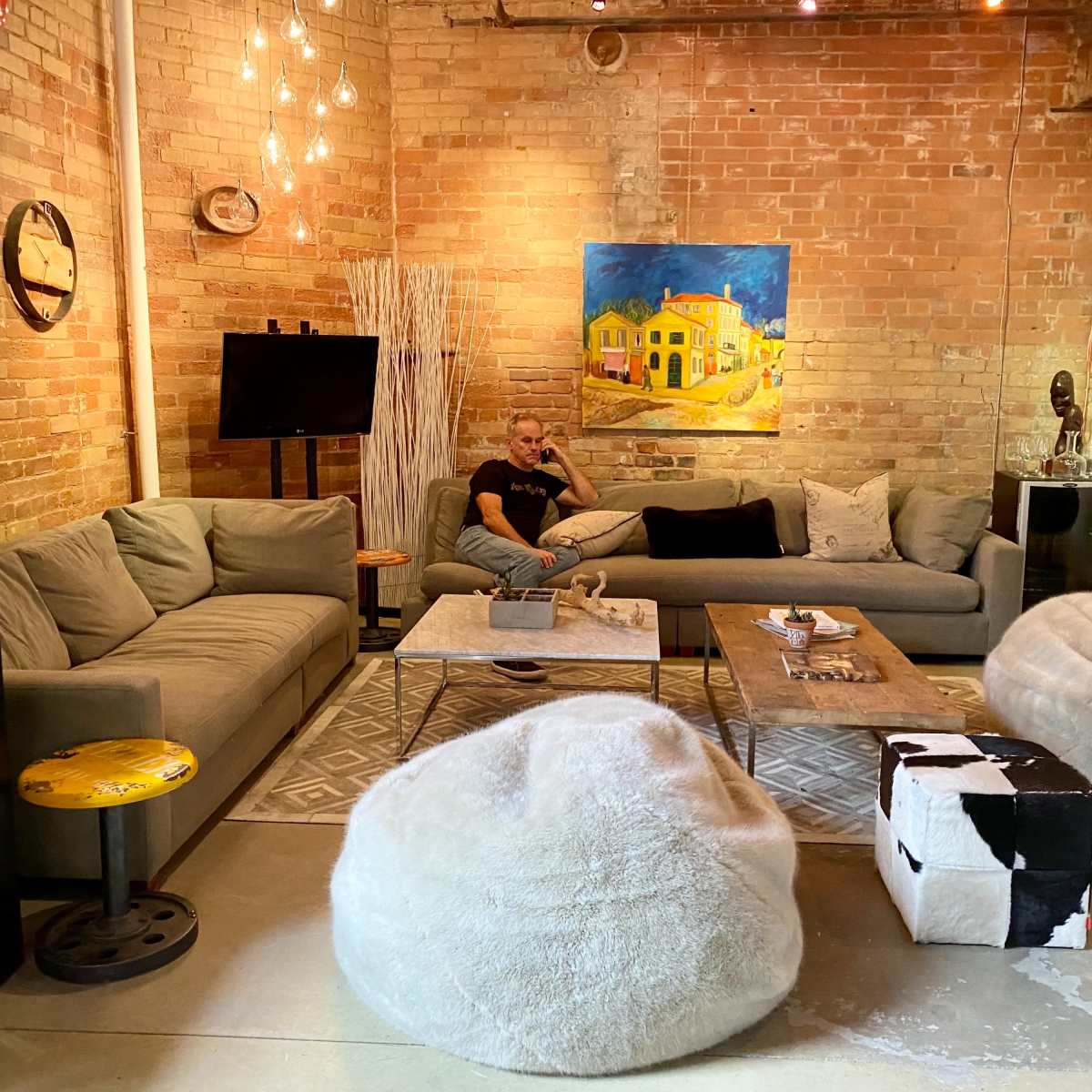A modern living room features a gray sectional sofa, a wooden coffee table, and bean bag chairs, with brick walls and artwork enhancing the decor.