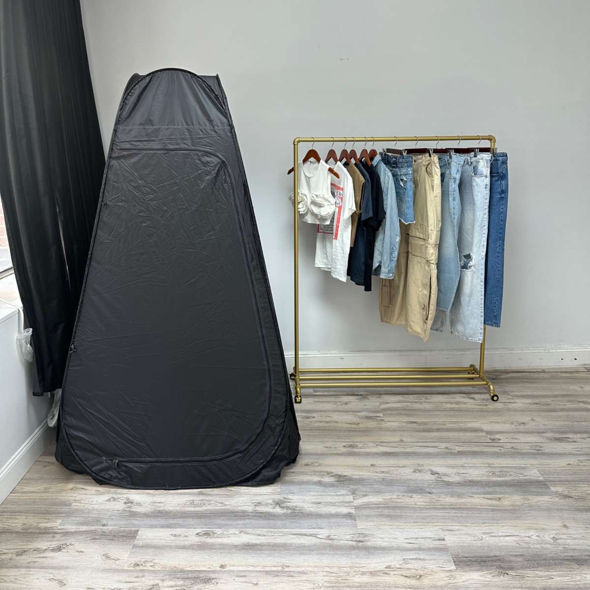 A black pop-up changing tent stands next to a gold clothing rack displaying various garments, including shirts and jeans, against a white wall.