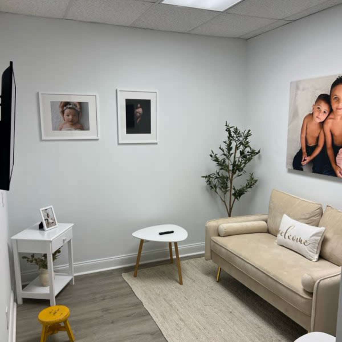 A small, minimalist living space with a beige couch, a white coffee table, and framed photographs on the wall, alongside a plant and a small yellow stool.