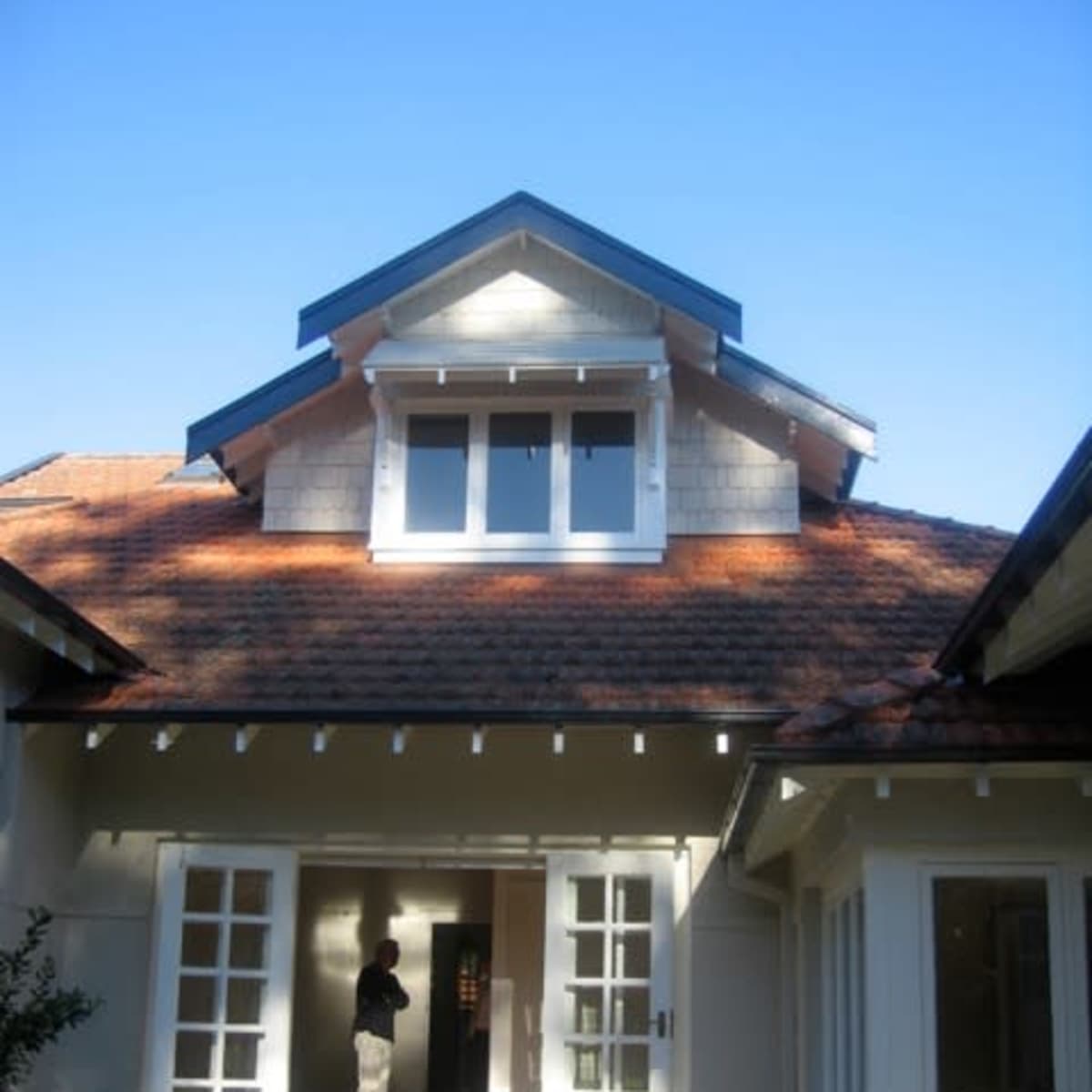 A person stands in the doorway of a house with a gabled roof and multiple windows under a clear blue sky.