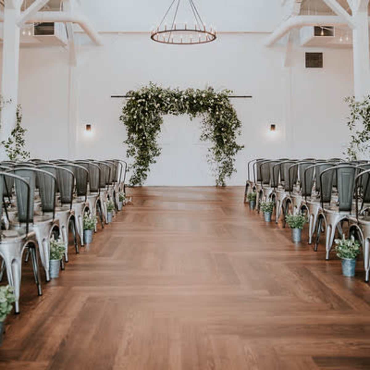 A spacious wedding venue with rows of metal chairs facing a greenery-adorned arch at the end of a wooden floor.