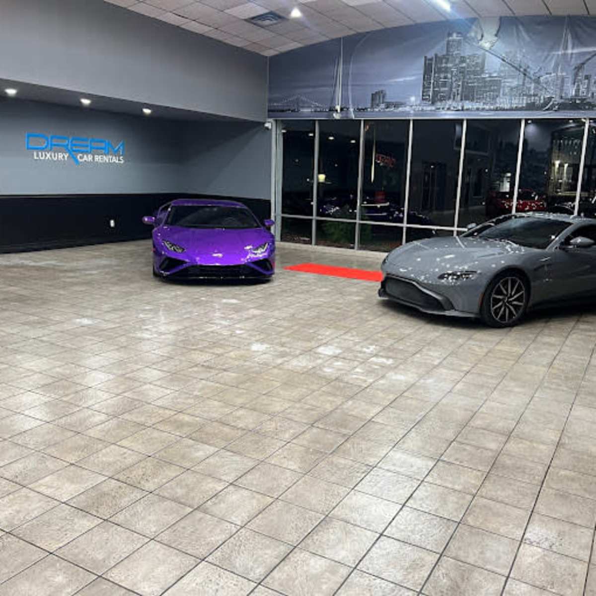 A car rental showroom featuring a purple sports car and a gray sports car parked on a tiled floor.