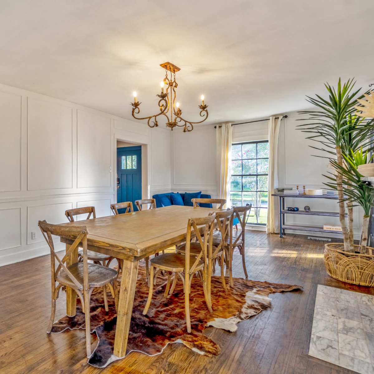 A spacious dining room features a large wooden table surrounded by chairs, a chandelier above, and a window offering natural light, with a doorway leading to another room.