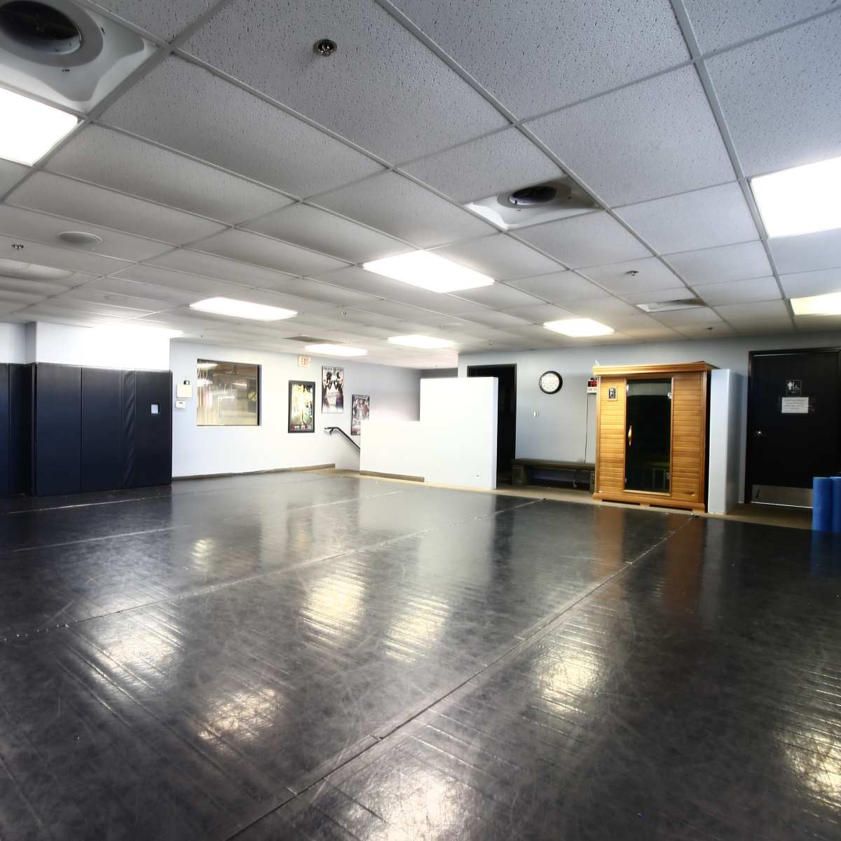A spacious, empty room with a black rubber flooring, light gray walls, and various wall-mounted features, including a clock and framed pictures.