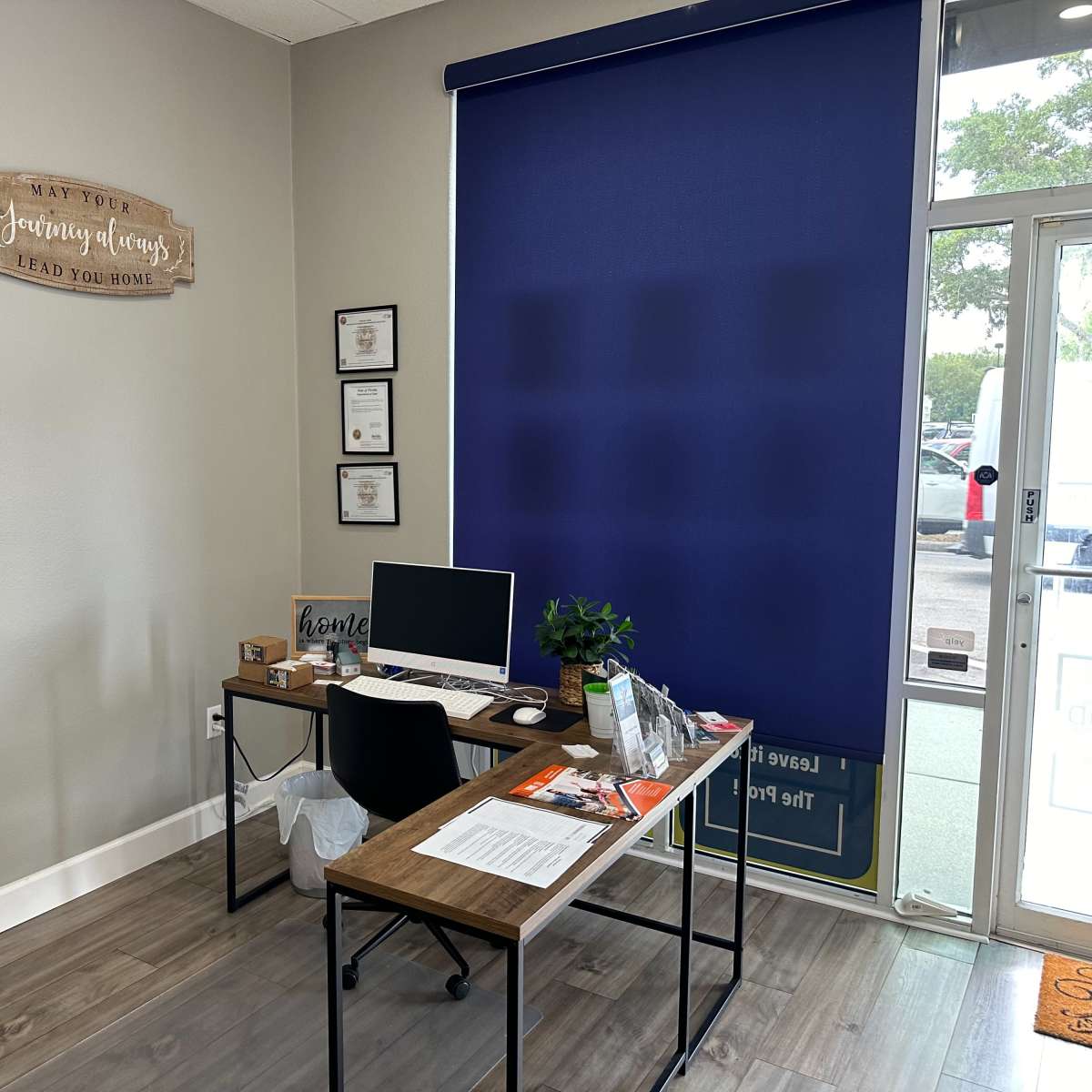 A small office space with a desk, a computer, and a blue window shade, accompanied by a decorative sign on the wall.
