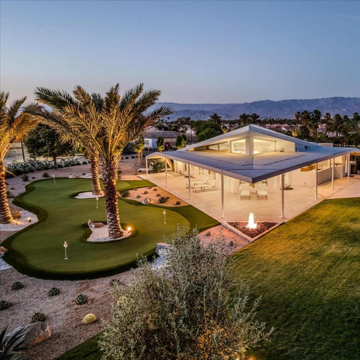 A modern house with a flat roof is situated amidst palm trees and landscaped green spaces, featuring an outdoor fire pit.