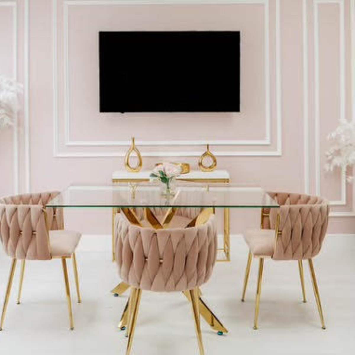 A modern dining room features a glass table surrounded by four blush pink chairs with gold legs, against a pale pink wall with a mounted television above.