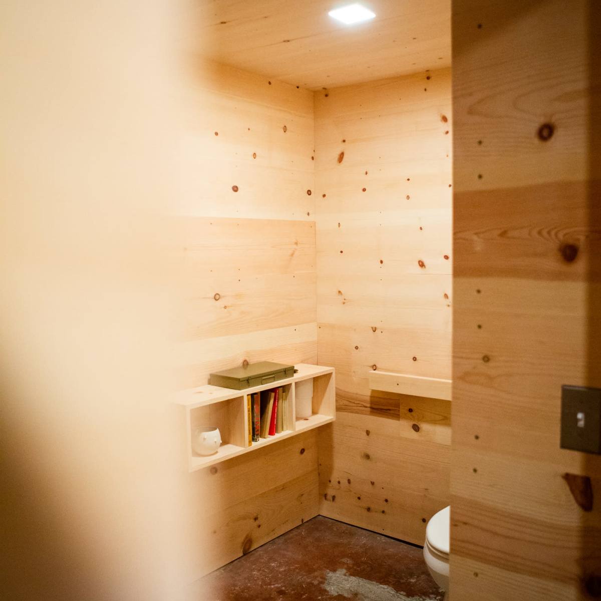 A minimalistic bathroom with wooden walls, a toilet, a small shelf containing books, and concrete flooring.