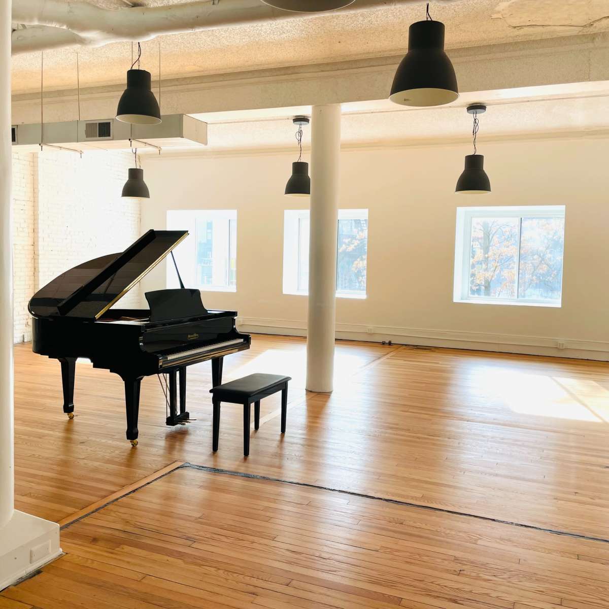A grand piano with a bench is positioned in the center of a spacious, well-lit room with hardwood floors and large windows.