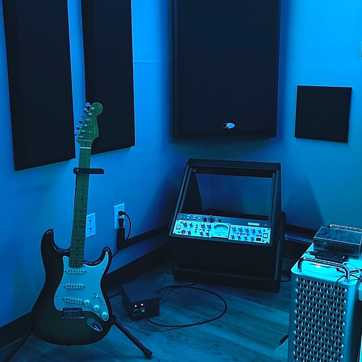 A music recording studio corner with an electric guitar, a speaker, a rack of audio equipment, and soundproofing panels on the walls.