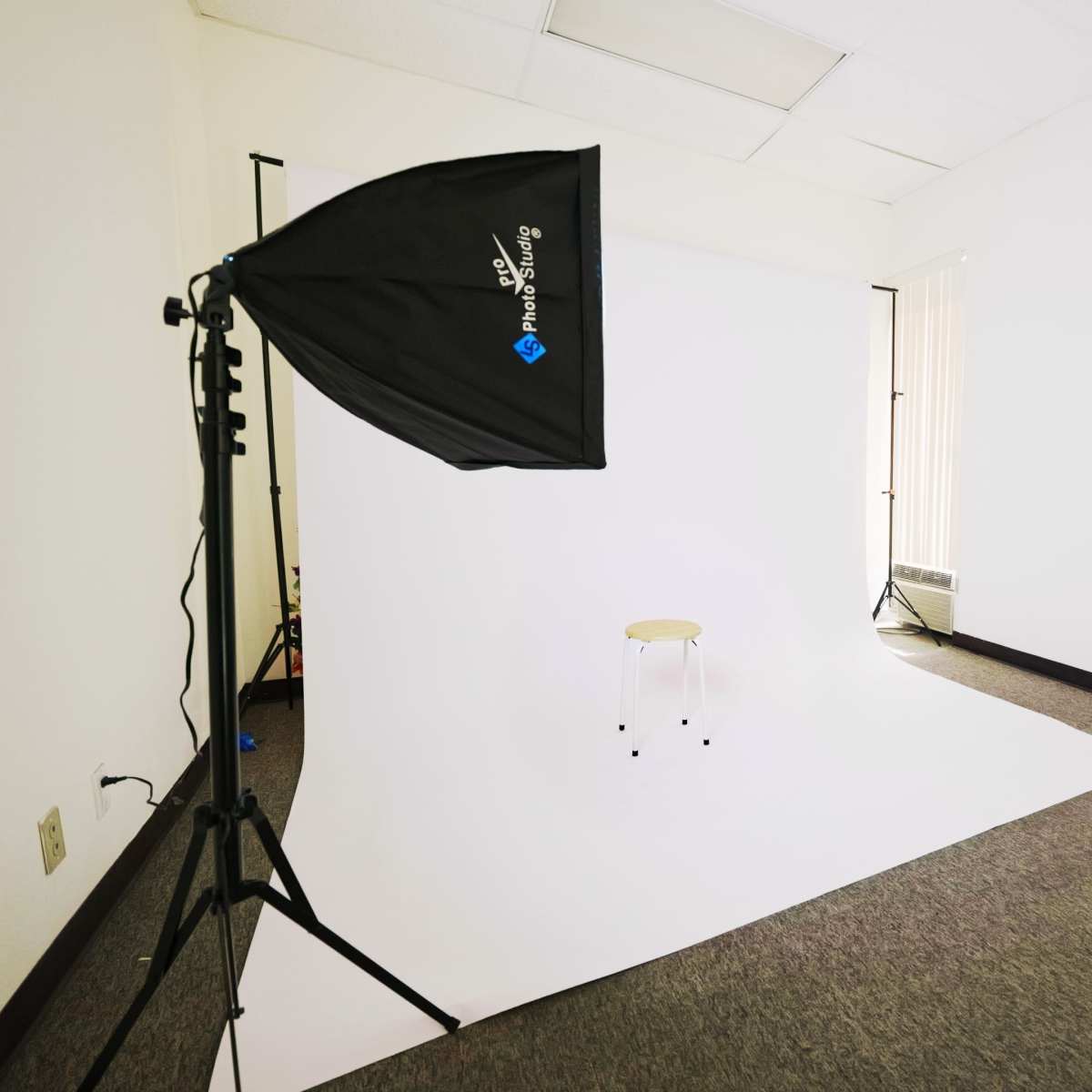 A photography studio is set up with two softbox lights facing a white backdrop and a single stool in the center.