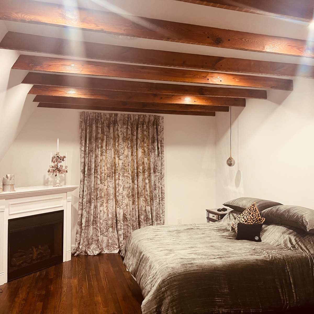 A bedroom features a large bed with gray bedding, rustic wooden beams overhead, and a window dressed with patterned curtains beside a decorative fireplace.