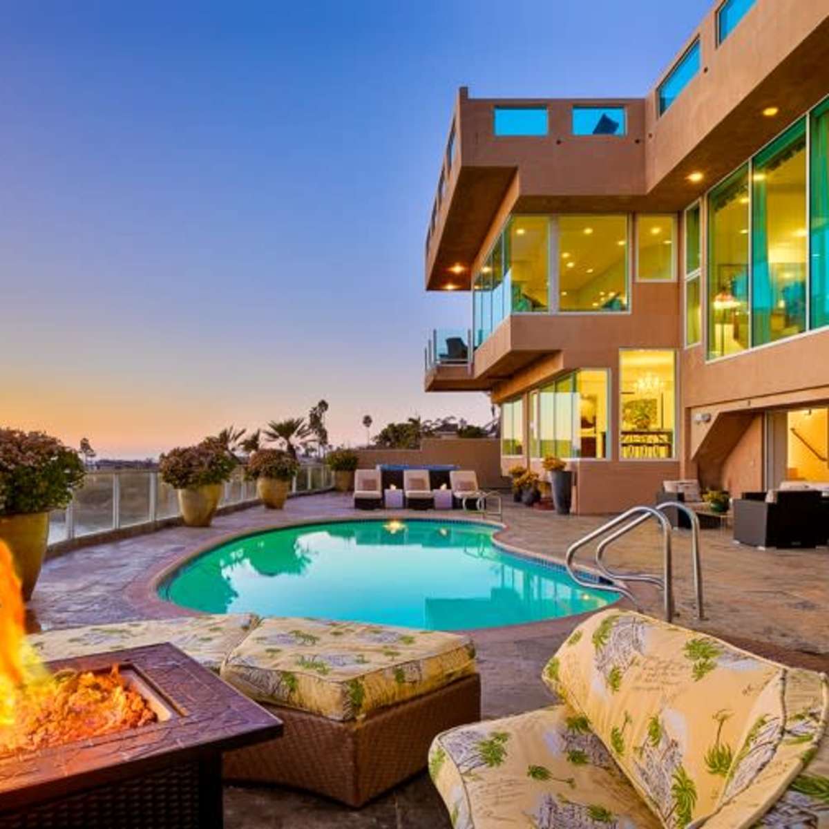 A modern home overlooks a pool with a fire pit, surrounded by seating, against a backdrop of a sunset over the ocean.
