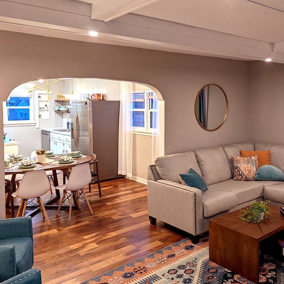 A modern living room with a gray sectional sofa, a wooden coffee table, and a view into a dining area featuring a round table and a kitchen.