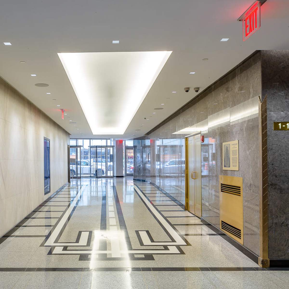 A modern hallway with polished floors, large framed artwork on the walls, and glass doors at the end leading to an outdoor area.