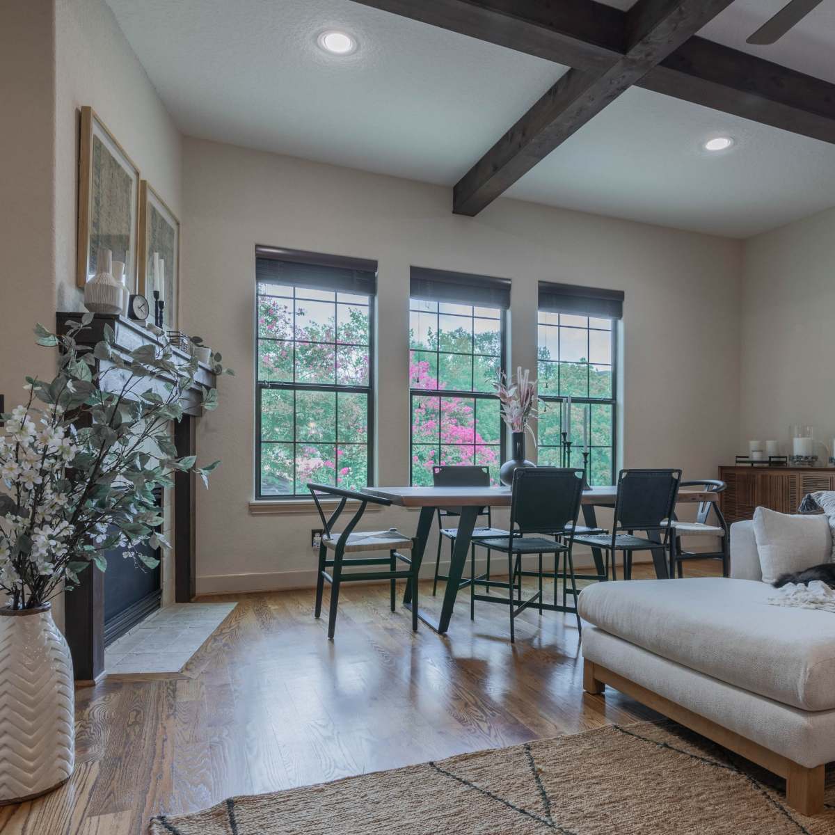 A modern living room with a beige sectional sofa, a dining table surrounded by black chairs, and large windows letting in natural light, framed by greenery outside.
