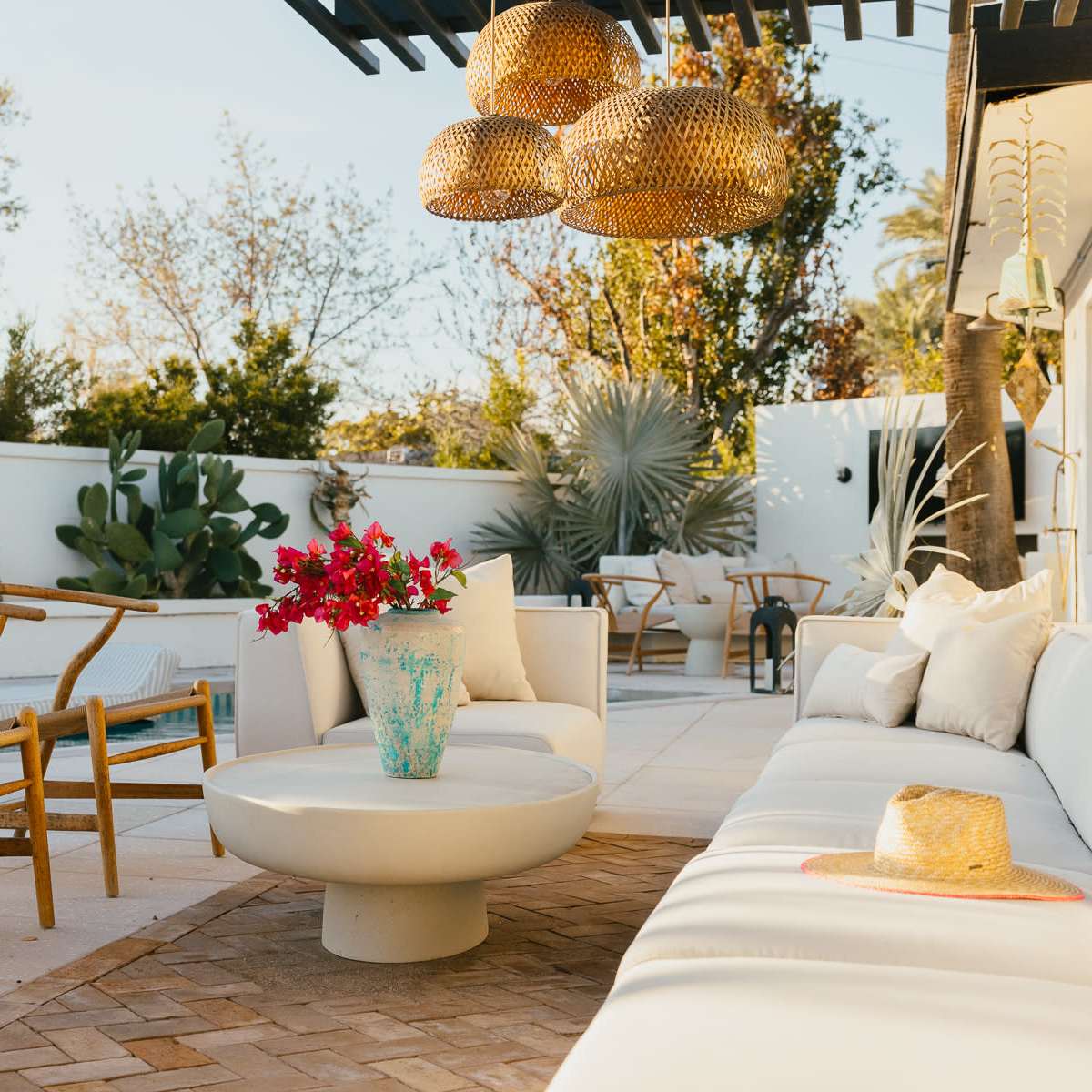The outdoor seating area features a white sofa, a round coffee table, and wooden chairs surrounded by various plants and a swimming pool in the background.