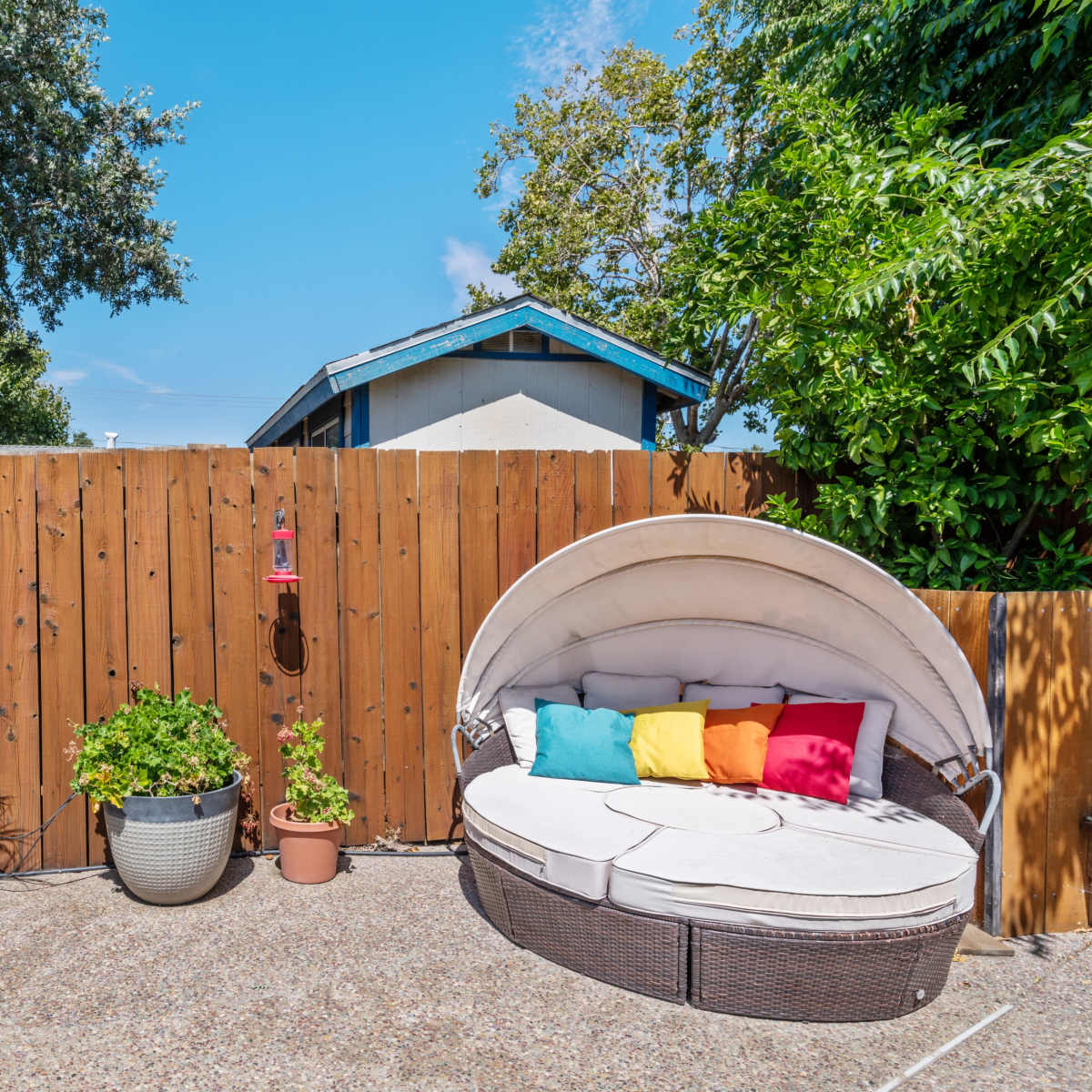 A circular outdoor daybed with colorful pillows is positioned on a patio surrounded by potted plants and a wooden fence.