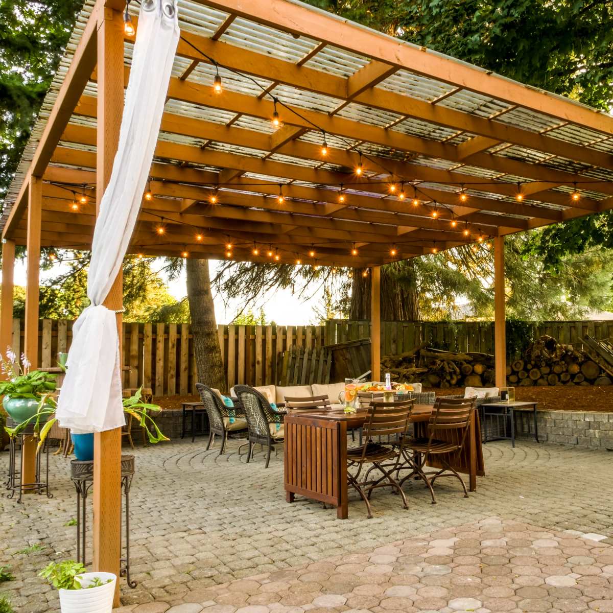 A wooden pergola with string lights over a brick patio featuring a dining table and chairs, surrounded by trees and a wooden fence.
