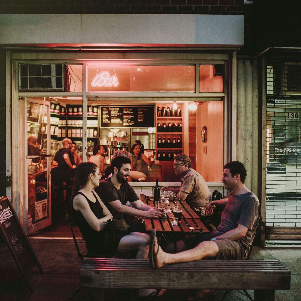 A group of four people sits at an outdoor table in front of a bar, with warm light spilling from inside where other patrons are visible.