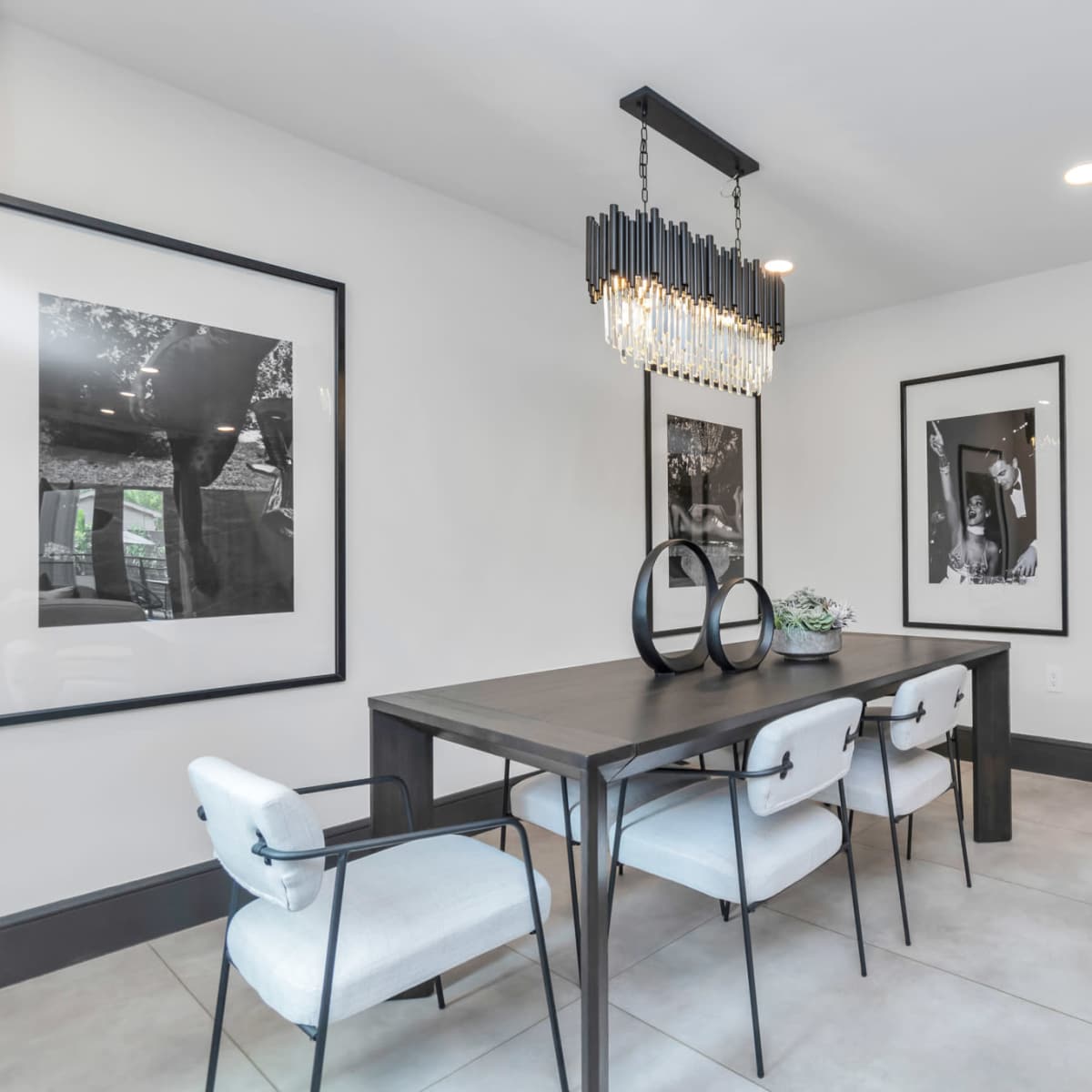A modern dining area features a rectangular table with white upholstered chairs, framed black-and-white photographs on the walls, and a stylish chandelier overhead.