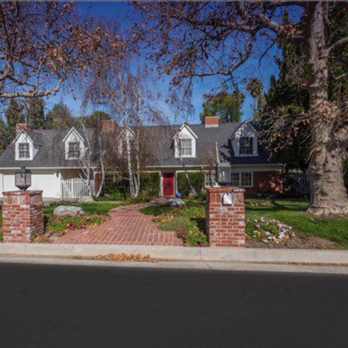 A two-story house with a sloped roof, multiple dormer windows, and a red front door is set behind a brick walkway and landscaped front yard.