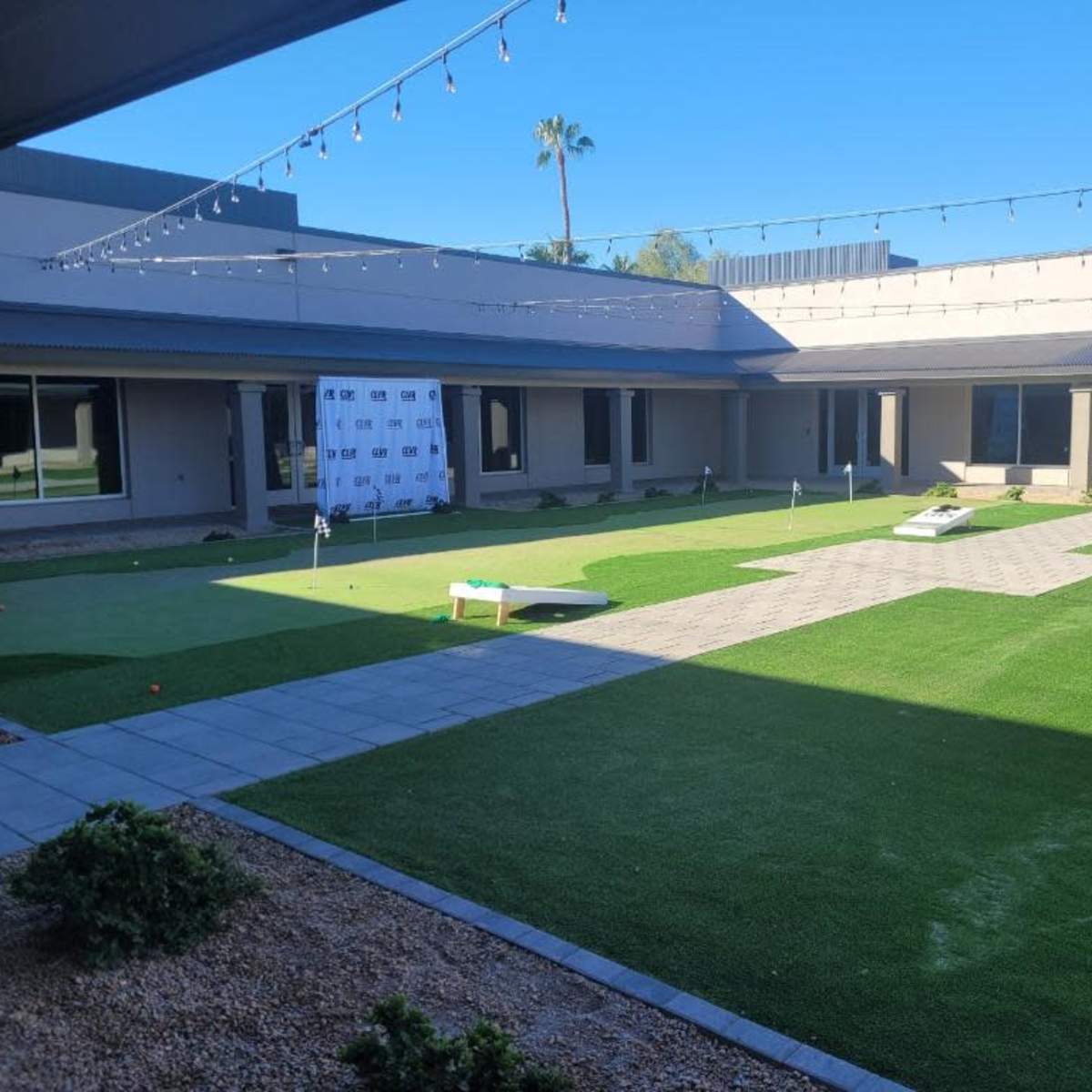 A landscaped courtyard featuring a green lawn, a paved pathway, and a backdrop with a logo, all surrounded by buildings.