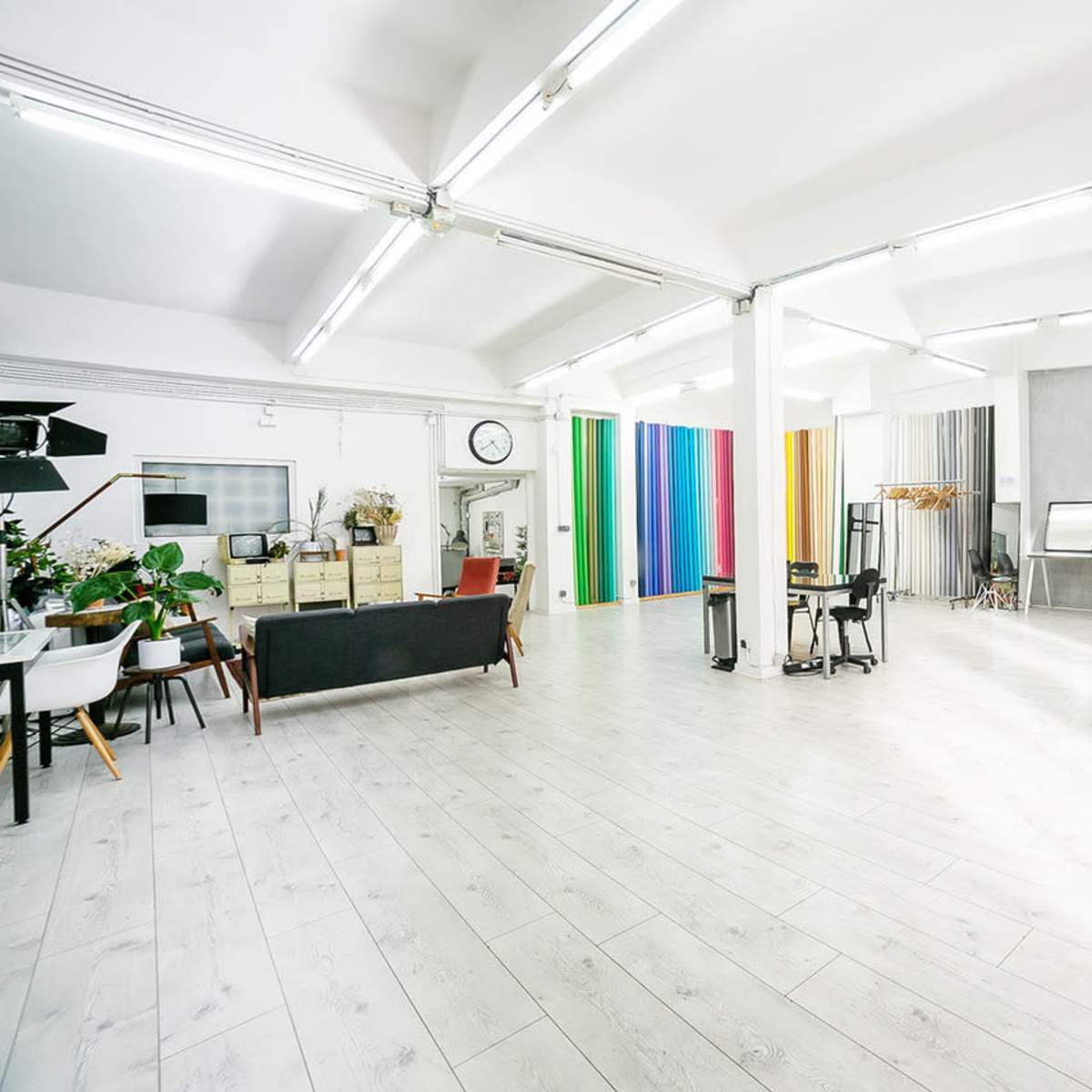 A spacious studio with light wooden flooring, various colored backdrop panels, and minimal furniture arranged in an open layout.