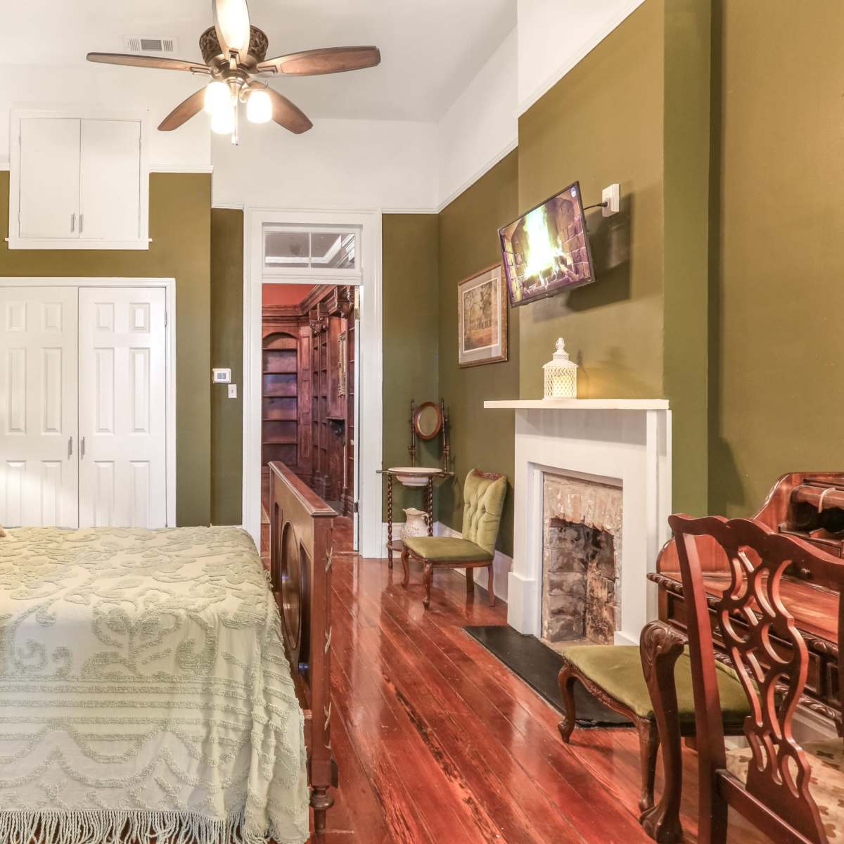 A bedroom features a wooden bed with a green bedspread, a decorative fireplace, and a ceiling fan, with walls painted in a dark green color.