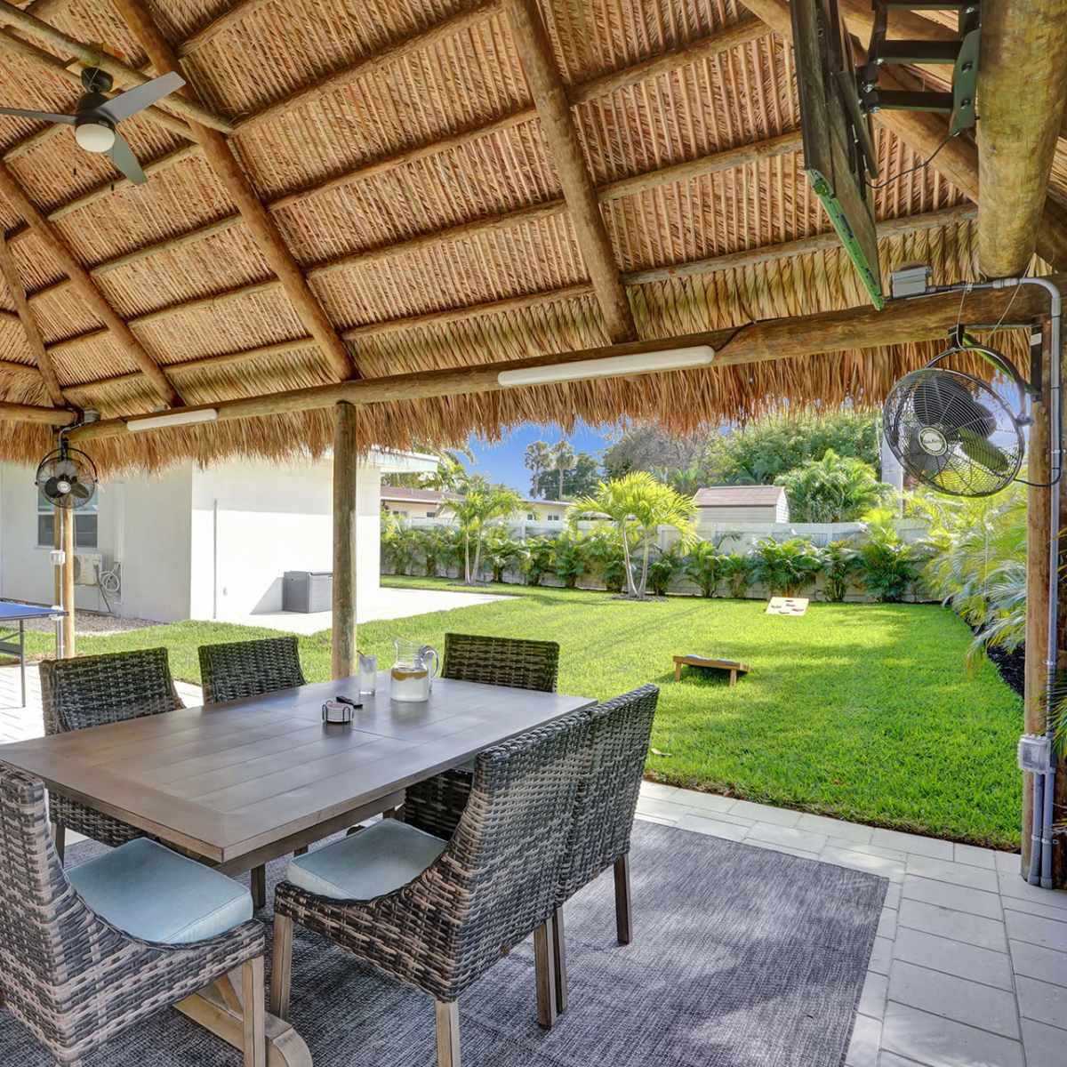 A shaded outdoor dining area with a table and chairs is set under a thatched roof, overlooking a grassy yard and a grill nearby.