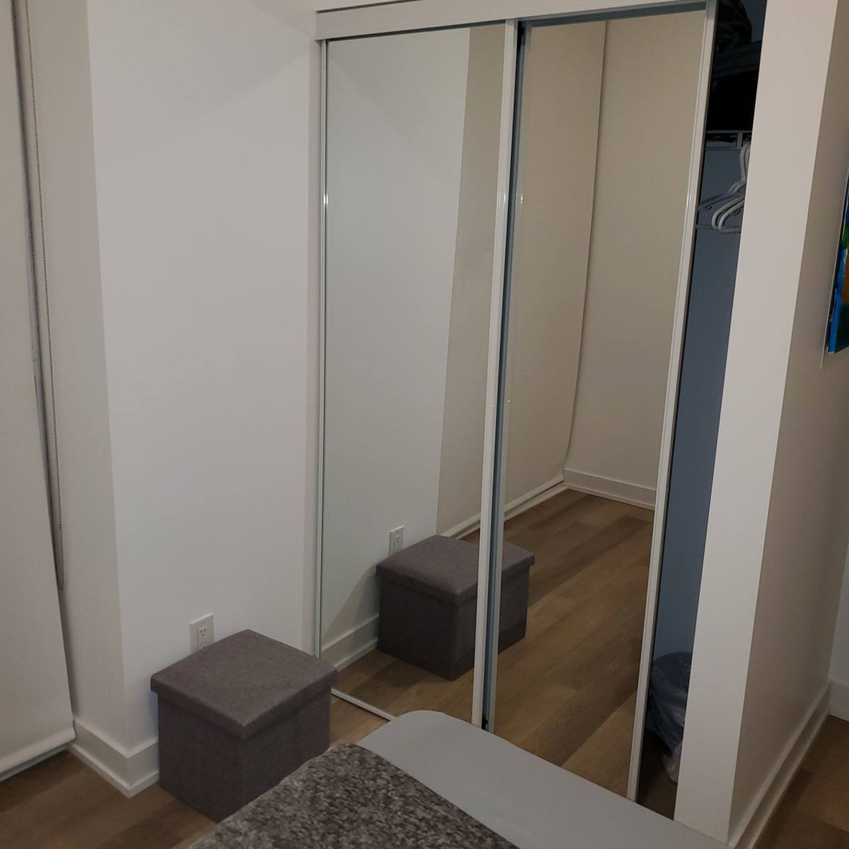 A bedroom with a bed in the foreground, a gray storage box near the foot of the bed, and a mirrored closet door against a light-colored wall.