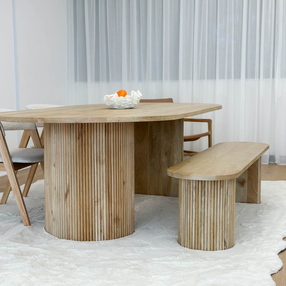 A wooden table with a curved surface and cylindrical legs is set in a light-filled room with a white rug and a bowl of fruit on top.