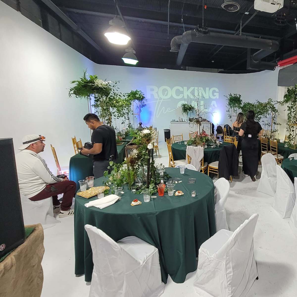 A spacious event venue features round tables set with green tablecloths, surrounded by guests mingling among decorative plants and a backdrop that reads "ROCKING THE V".