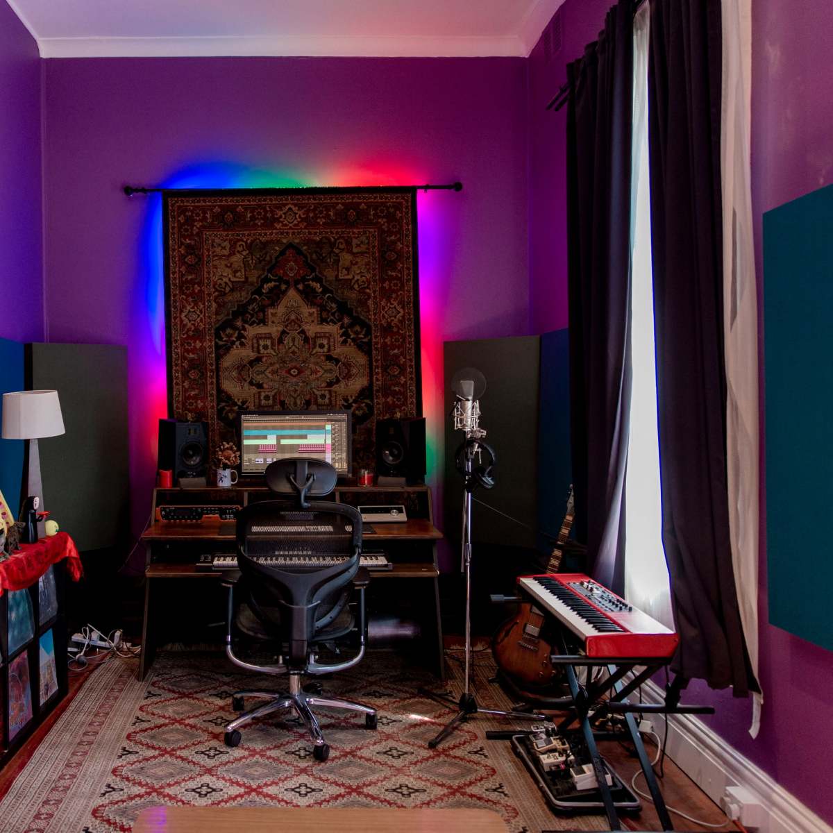 A music production studio features a desk with a computer, a microphone, and colorful soundproofing panels on the walls.