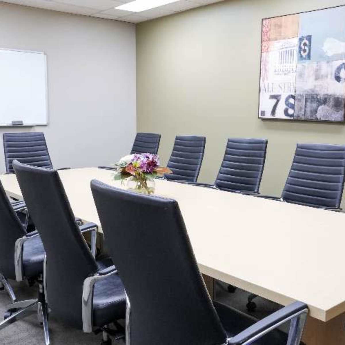 A conference room features a long table surrounded by eight black leather chairs and a floral centerpiece, with a whiteboard on one wall.