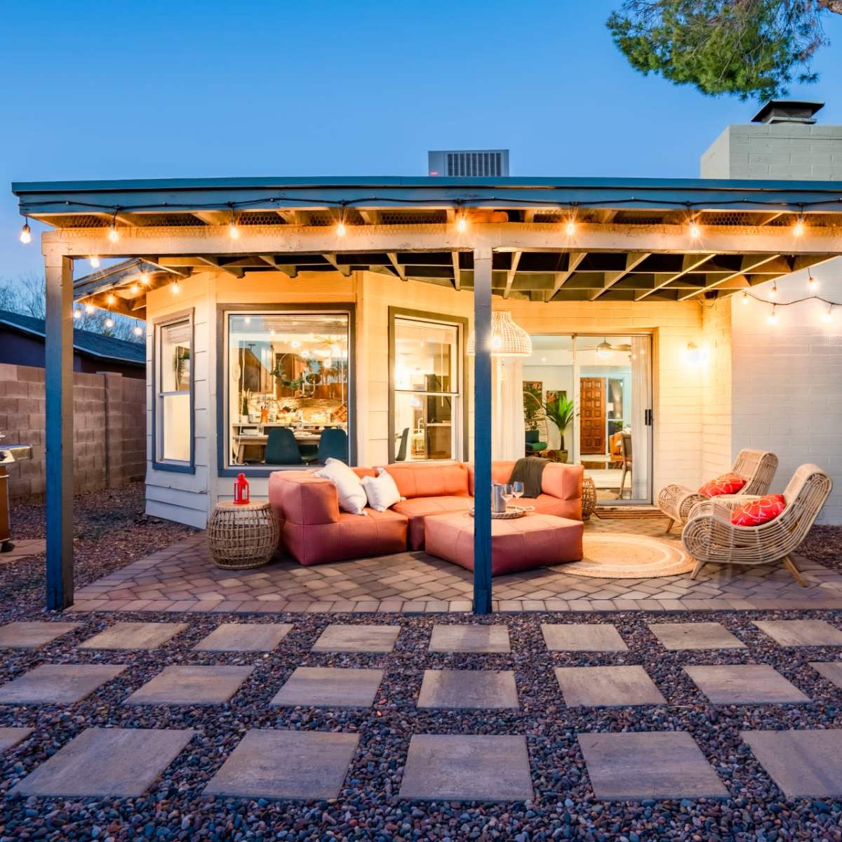 A cozy outdoor patio area with a sectional sofa, two chairs, and a grill, surrounded by landscaping and illuminated by string lights.