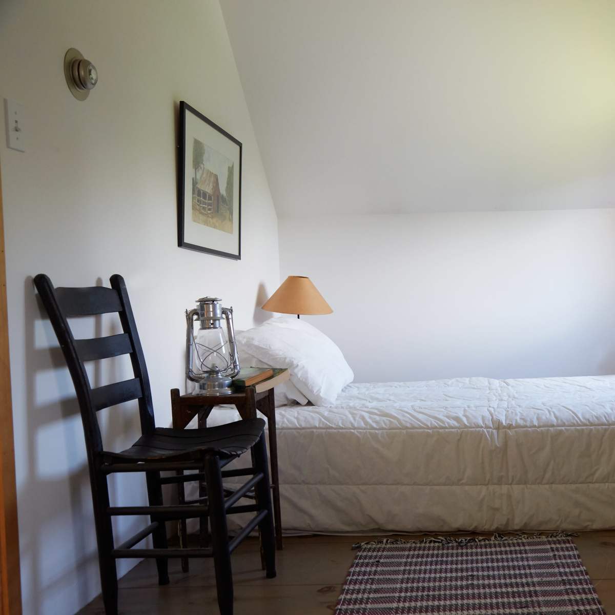 A simple bedroom featuring a single bed, a chair, a small side table with a lantern, and a vintage dresser.