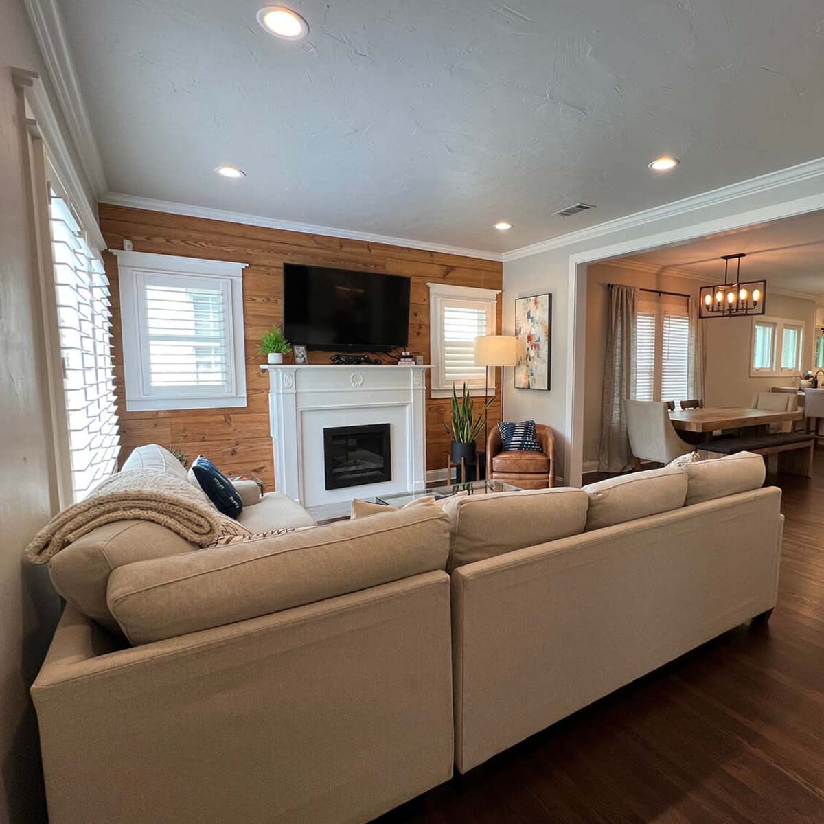 A beige sofa is positioned in a living room that features a fireplace and a wall-mounted TV, with a view into a dining area and kitchen beyond.