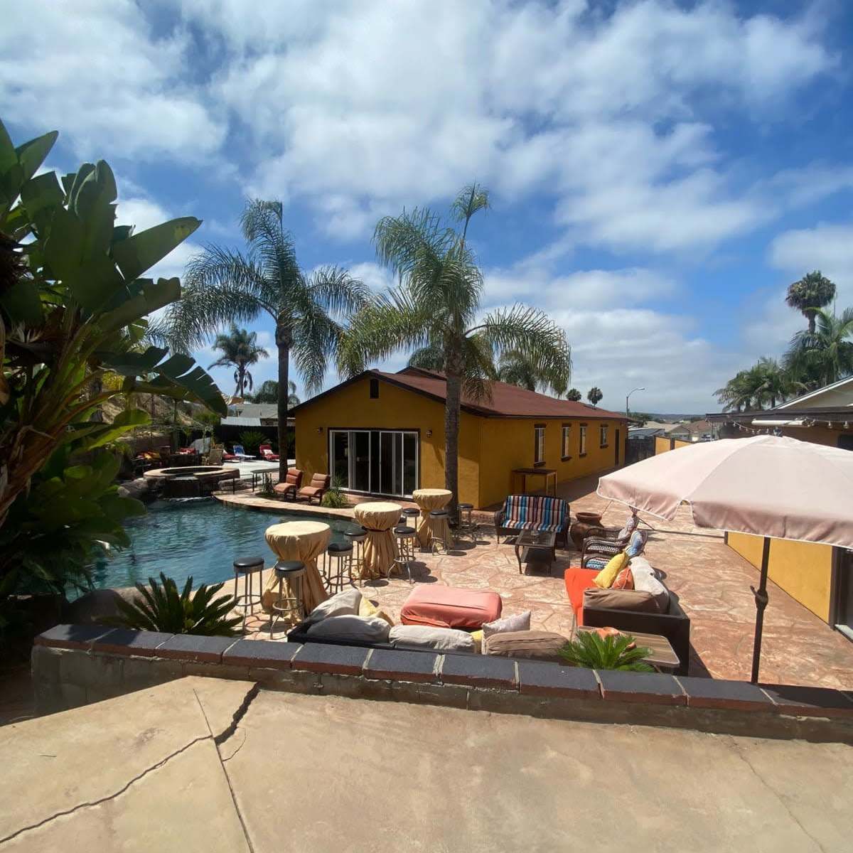 A sunny outdoor pool area with palm trees, lounge chairs, and a yellow building in the background.