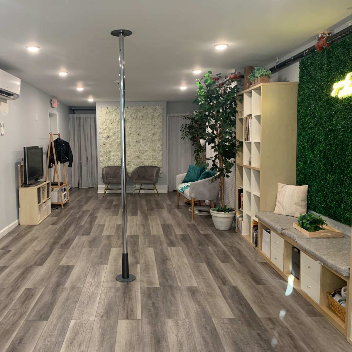 A spacious indoor studio or room featuring a pole in the center, furnished with chairs, a TV, shelves, and decorative plants against a light gray wall.