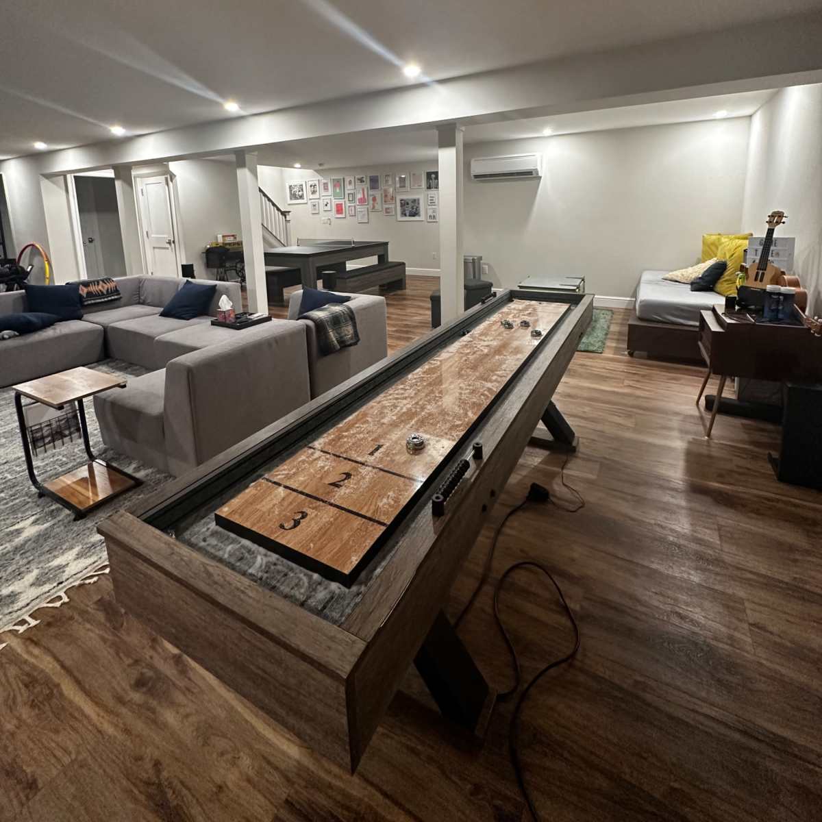 A shuffleboard table is positioned in the center of a modern, dimly lit basement lounge that features various seating areas and wall art.