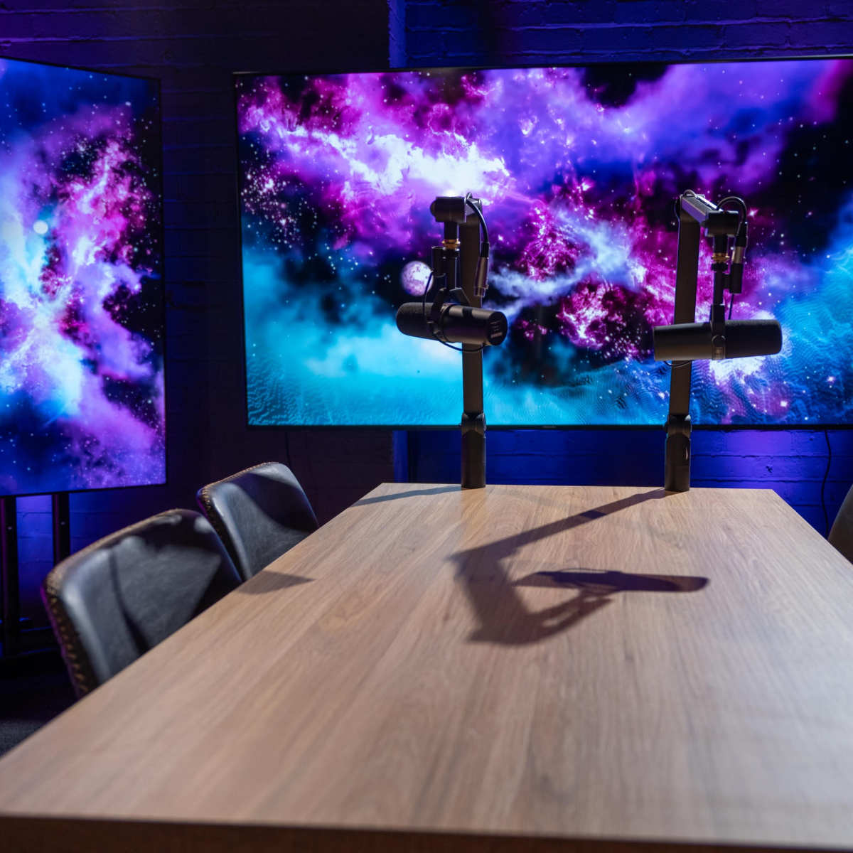 A wooden conference table is positioned in front of two large screens displaying colorful cosmic visuals in a dimly lit room.