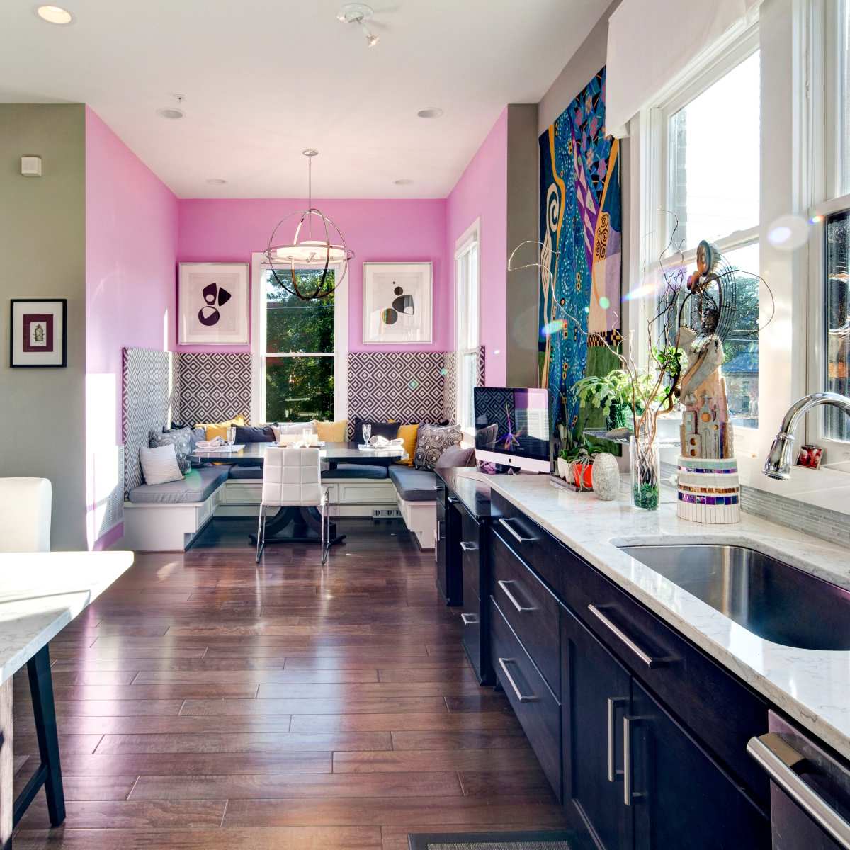 A modern kitchen with a marble countertop, dark cabinetry, and a dining area featuring a pink wall and a seating nook by large windows.