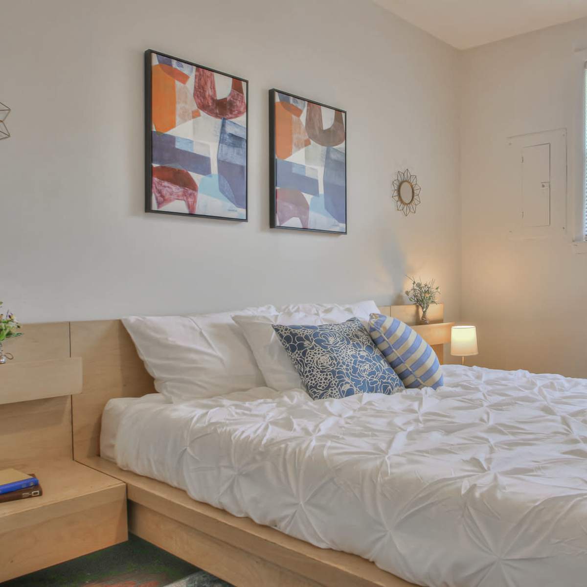 A neatly arranged bedroom features a double bed with white bedding, two framed abstract paintings on the wall, and a small nightstand with a lamp.