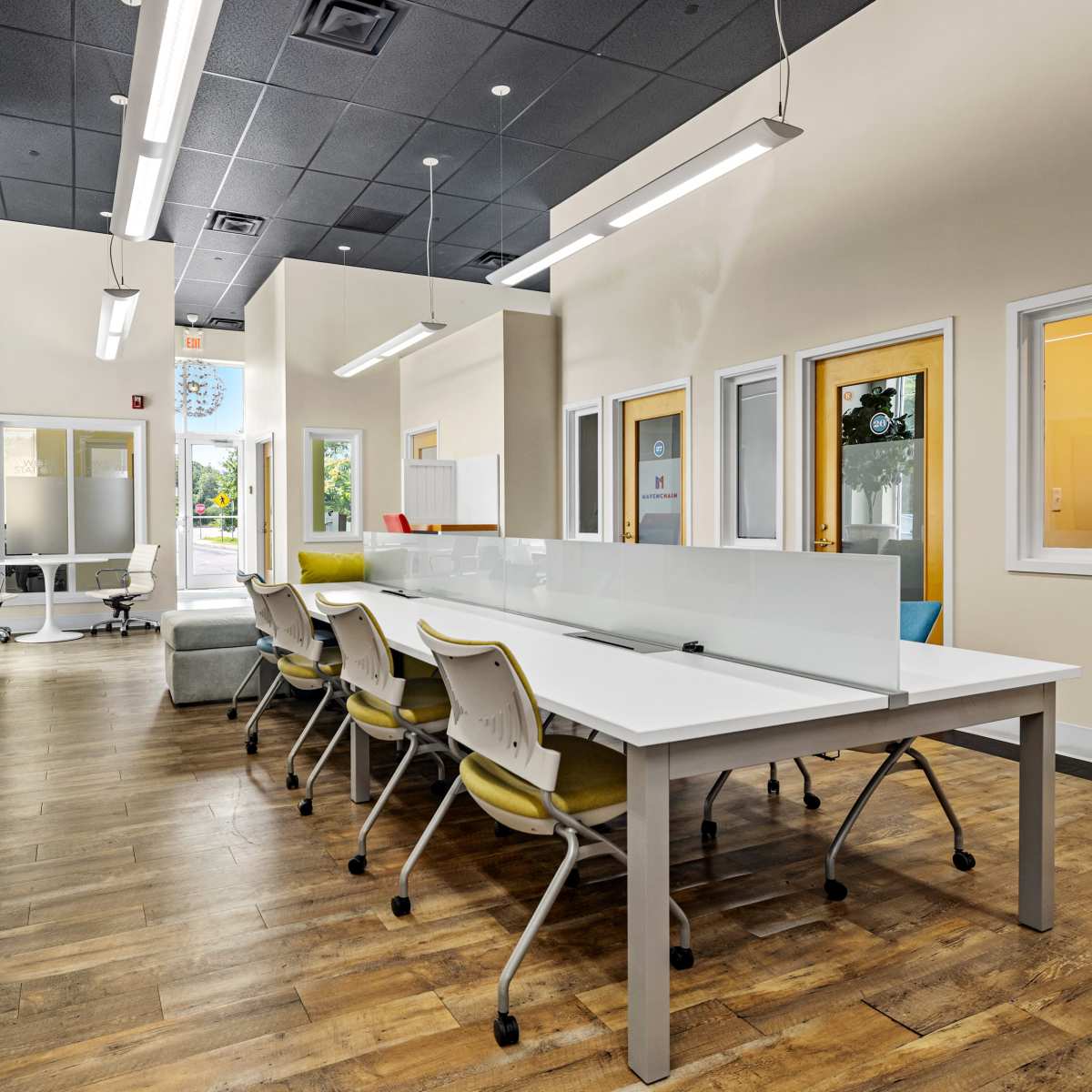 A modern office space with a large white table surrounded by yellow chairs, wooden flooring, and large windows providing natural light.