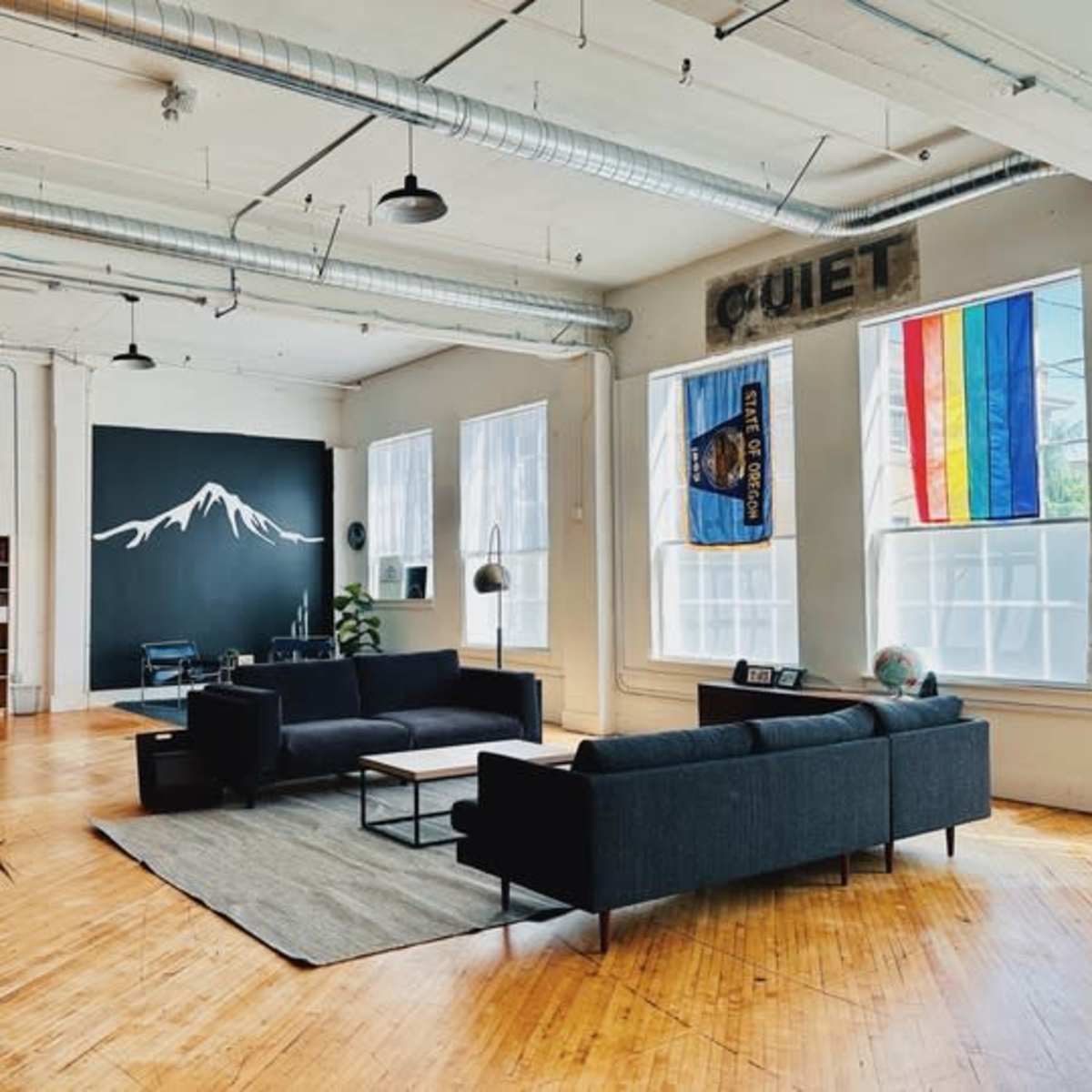 A spacious, modern interior with two dark sofas facing a large wall featuring a mountain mural and colorful flags hanging in the windows.