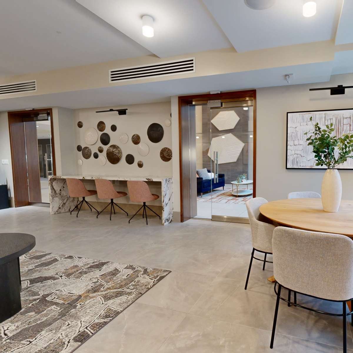 A modern interior space featuring a round dining table surrounded by upholstered chairs, a patterned rug, and decorative wall art, with large windows providing natural light.